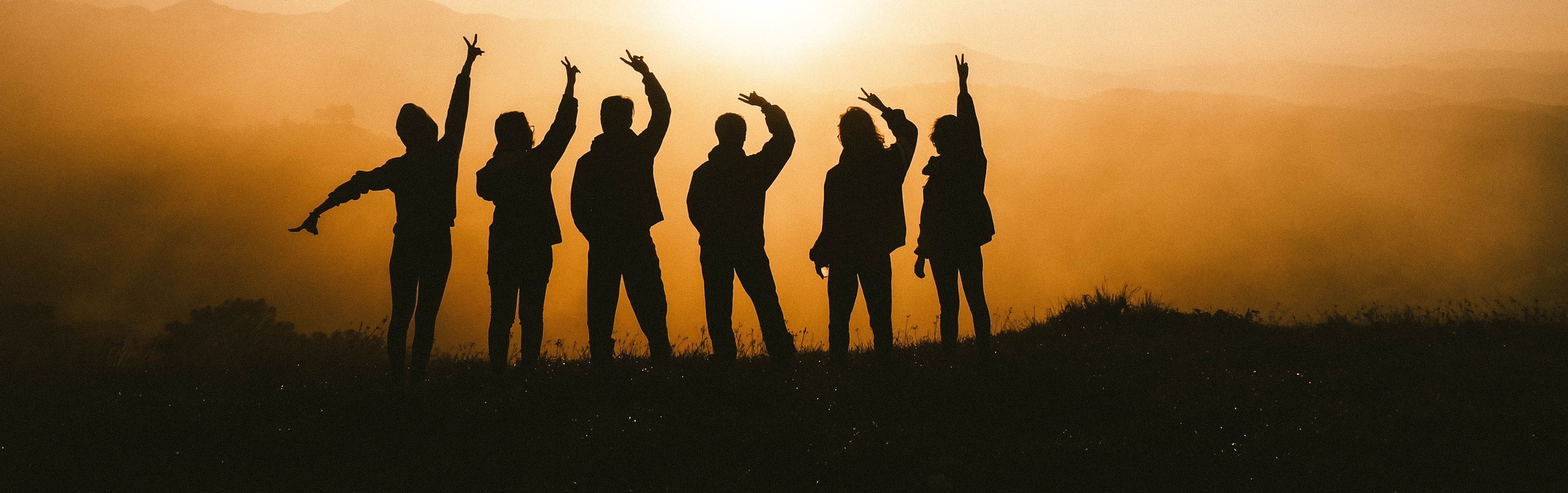 Silhouette of a group of friends with arms raised atop a mountain peak during sunset